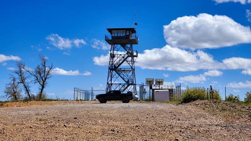 Lookout Tower enclosed with chain link gate. Black truck parked parallel to tower. Large white clouds in blue sky.